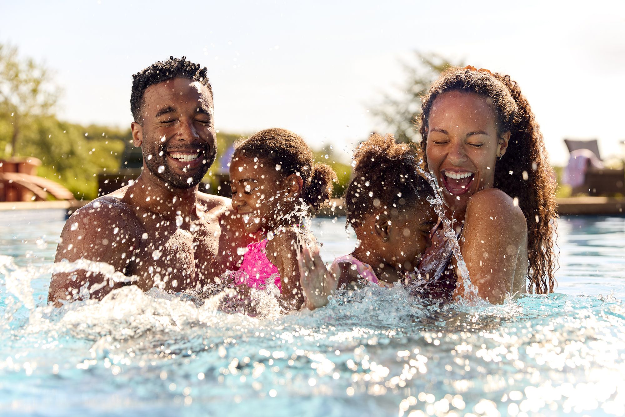 A family of four, including two little girls and their parents, in a swimming pool with their eyes closed as water splashes on them.